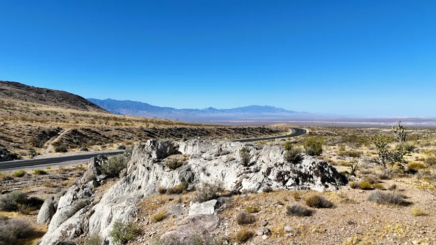 Expansive desert landscape with a paved road curving through rolling terrain, featuring distant mountains under a clear blue sky