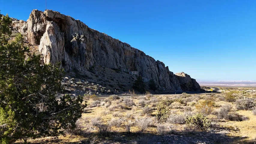 Natural rock formation bordering a winding road, with scattered desert flora and distant plains under a clear sky