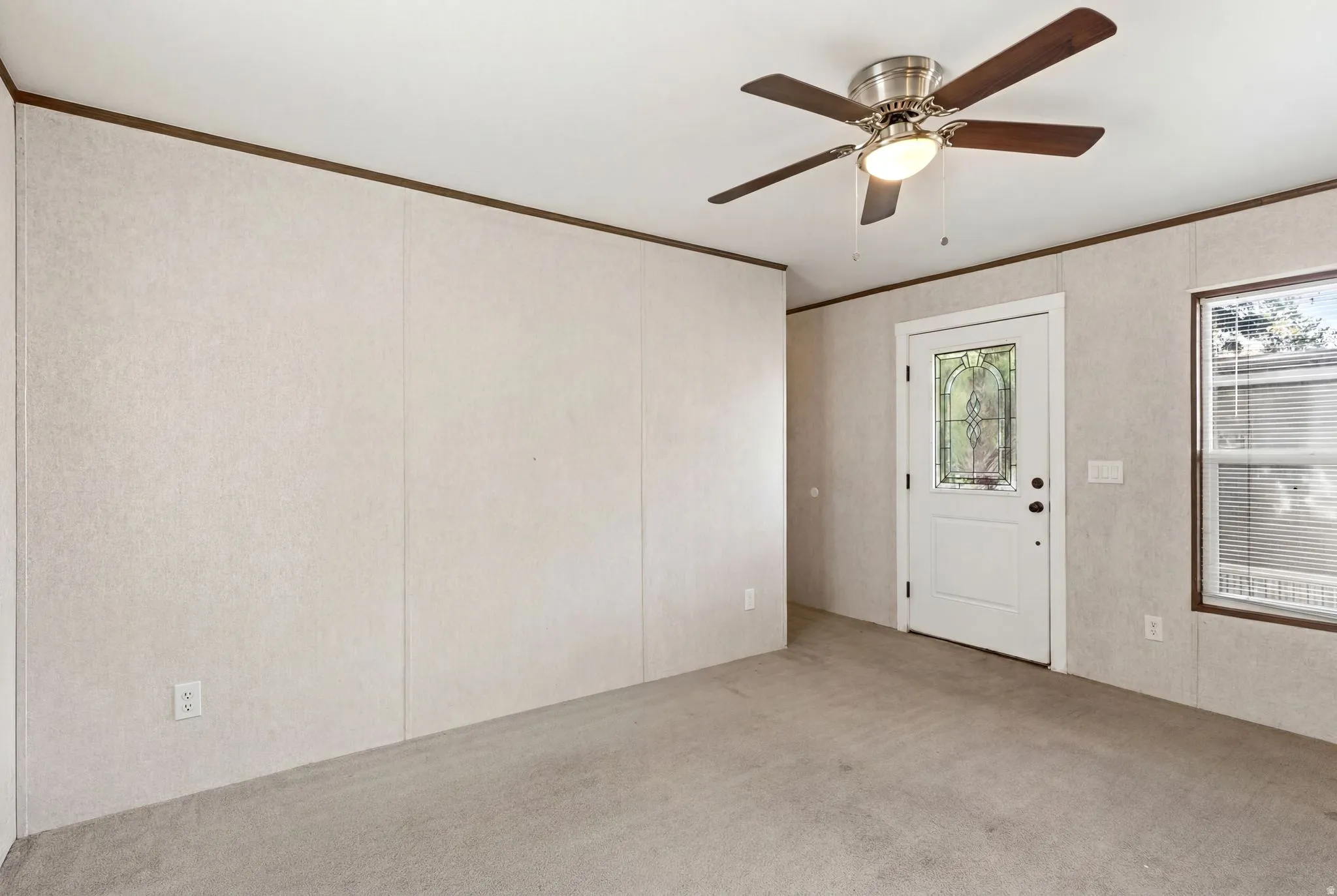 Neutral-toned panel walls with wood trim, featuring a ceiling fan with light fixture, a white door with decorative glass insert, and a window with blinds