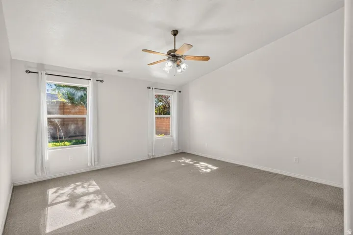 Spacious room featuring a vaulted ceiling with a wood-finish ceiling fan, two large windows, light gray carpet, and white walls