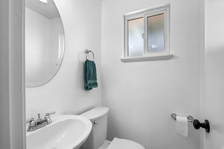 Powder room featuring a circular mirror, a ceramic pedestal sink with a chrome faucet, a white toilet, and a frosted window with white trim