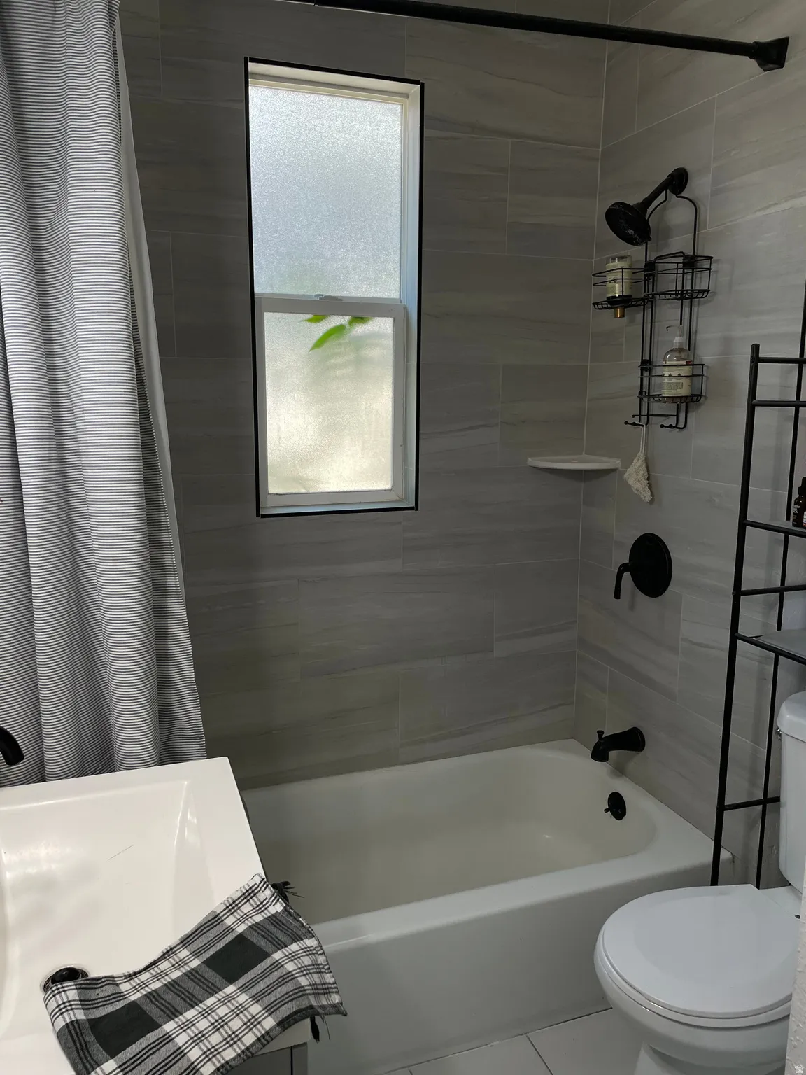 Bathroom featuring light gray tiled shower surround, a white bathtub, black fixtures, and a rectangular frosted window