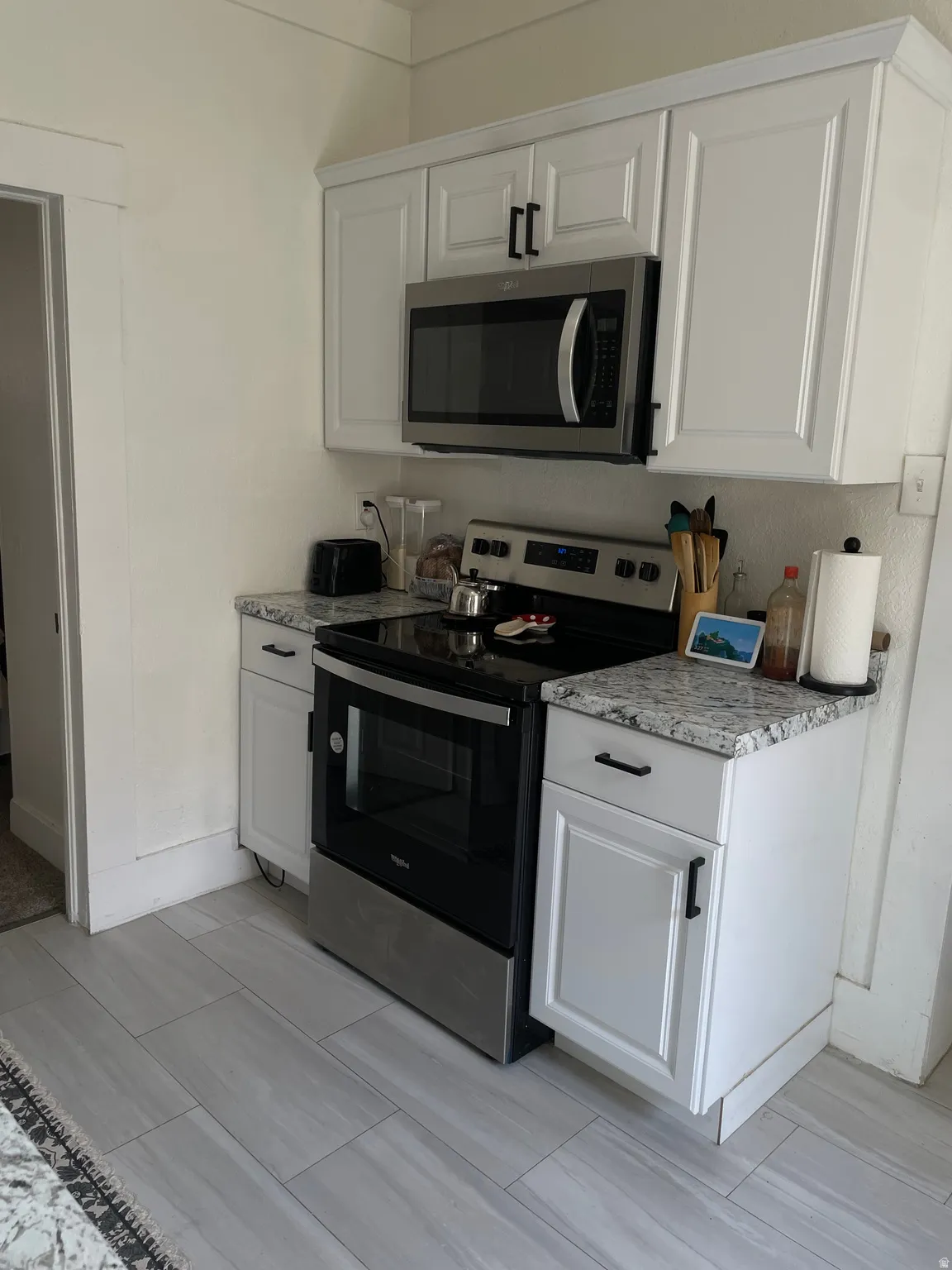 Kitchen area featuring white cabinetry with dark hardware, a stainless steel microwave, a stainless steel range, granite-style countertops, and light-toned tile flooring