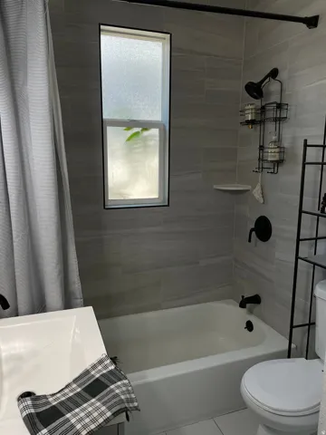 Bathroom featuring light gray tiled shower surround, a white bathtub, black fixtures, and a rectangular frosted window