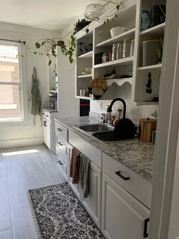 Kitchen featuring a double basin stainless steel sink, solid surface countertop, white shaker-style cabinetry with matte black hardware, integrated open shelving, and light wood-finish flooring