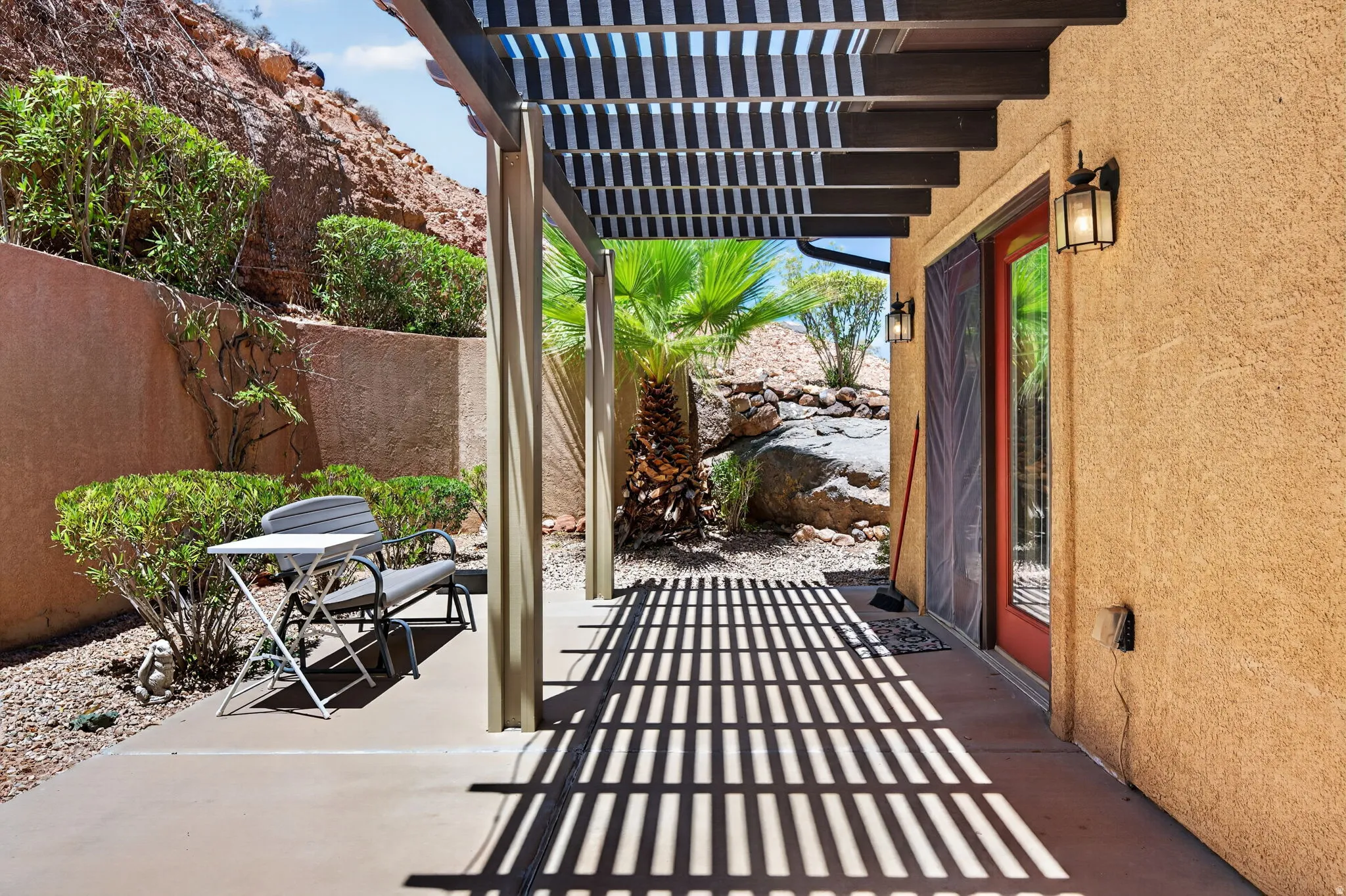 Shaded patio featuring a wood-beam pergola, textured stucco exterior, and a red-framed glass door