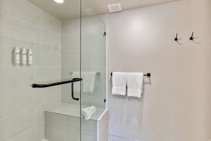 Glass-enclosed shower featuring light grey rectangular wall tiles, a built-in shower bench, a black shower handle, and a black grab bar