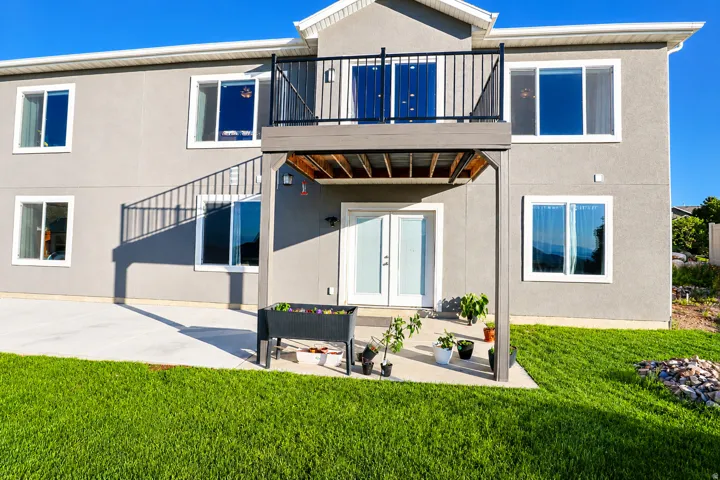 Rear elevation featuring a stucco finish, multiple windows, a second-story balcony with black metal railing, and a ground-level patio with double French doors