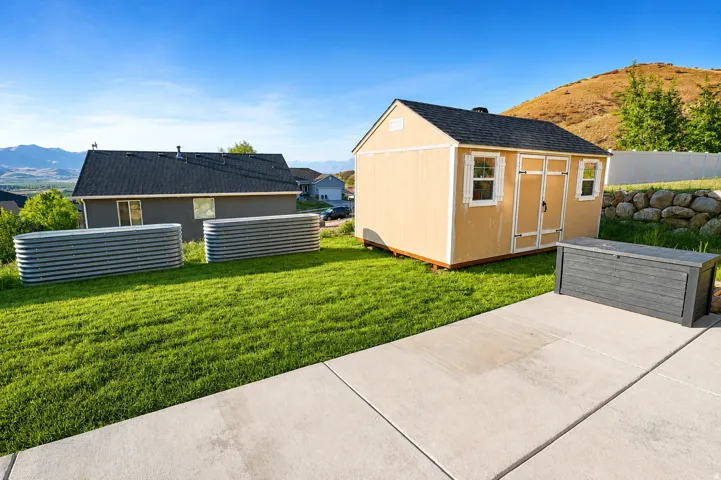 Spacious backyard featuring a concrete patio, a detached shed with a shingled roof, two galvanized steel raised garden beds, a sloped rock retaining wall, and a distant mountain backdrop