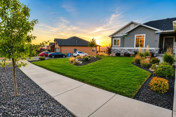 Contemporary gray stucco exterior featuring a dark gray shingle roof, stone accenting, a concrete walkway, and a meticulously maintained lawn with varied landscaping