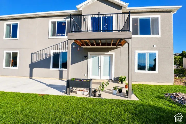 Rear elevation featuring a stucco finish, multiple windows, a second-story balcony with black metal railing, and a ground-level patio with double French doors
