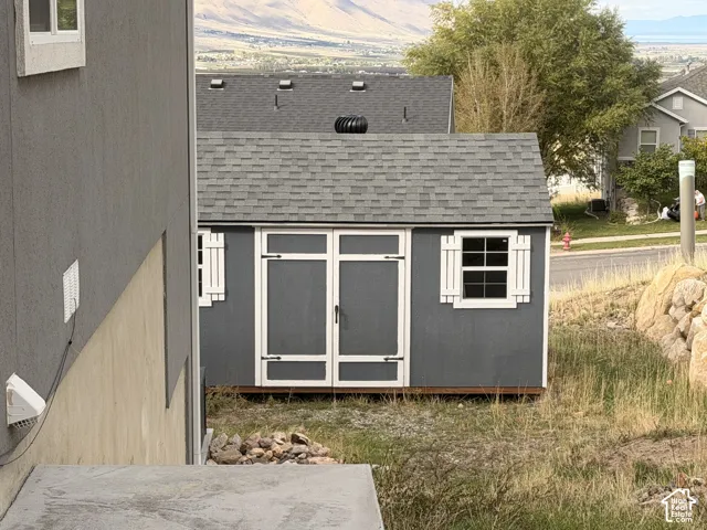 Exterior storage shed featuring a grey finish, shingle roof, double doors, and white shutter accents