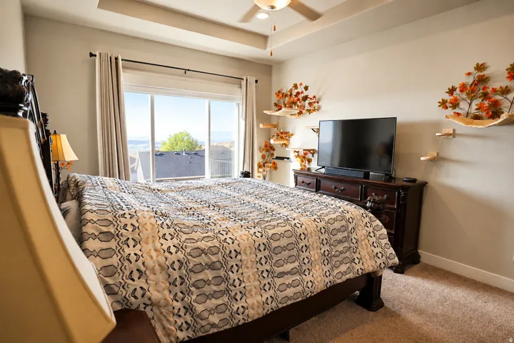 Carpeted bedroom featuring a tray ceiling with a fan light fixture, sliding glass doors with drapery, and neutral wall tones