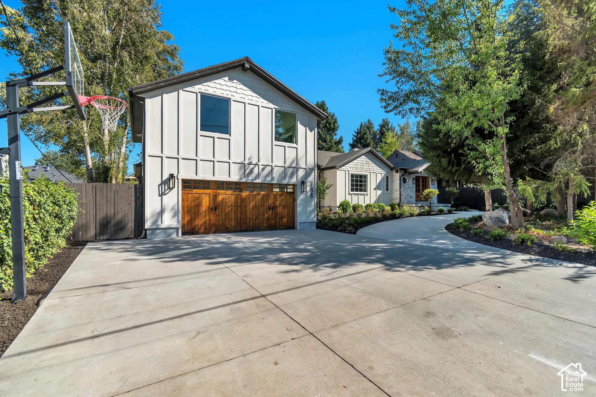 Expansive concrete driveway and contemporary exterior featuring a light-colored facade with vertical and horizontal trim, a two-car wood-finish garage door, and a basketball hoop