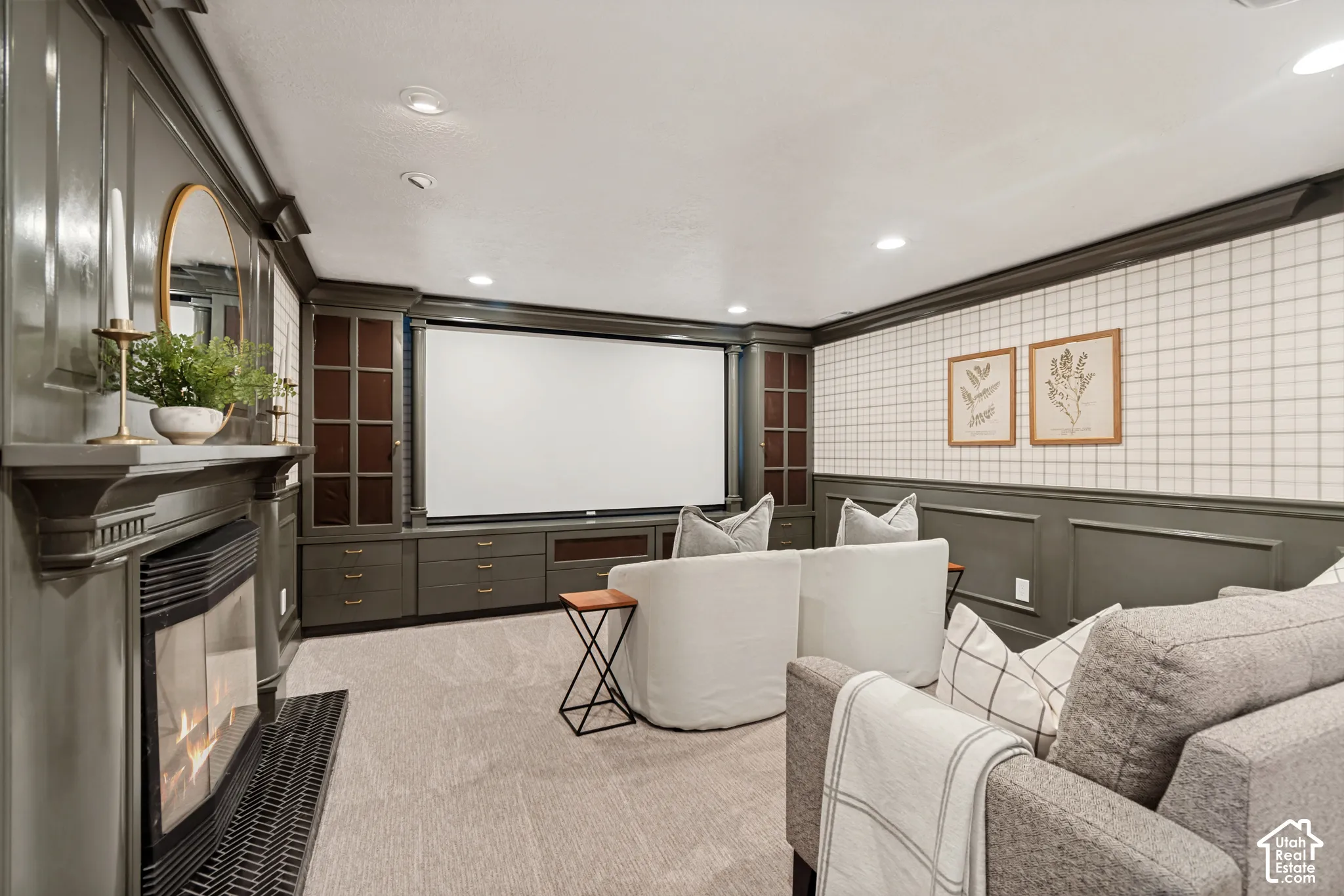 Integrated media room featuring a large projector screen, built-in cabinetry with glass-front display cases and drawer storage, a fireplace with a mantel, recessed ceiling lighting, and wainscoting with a patterned wall covering