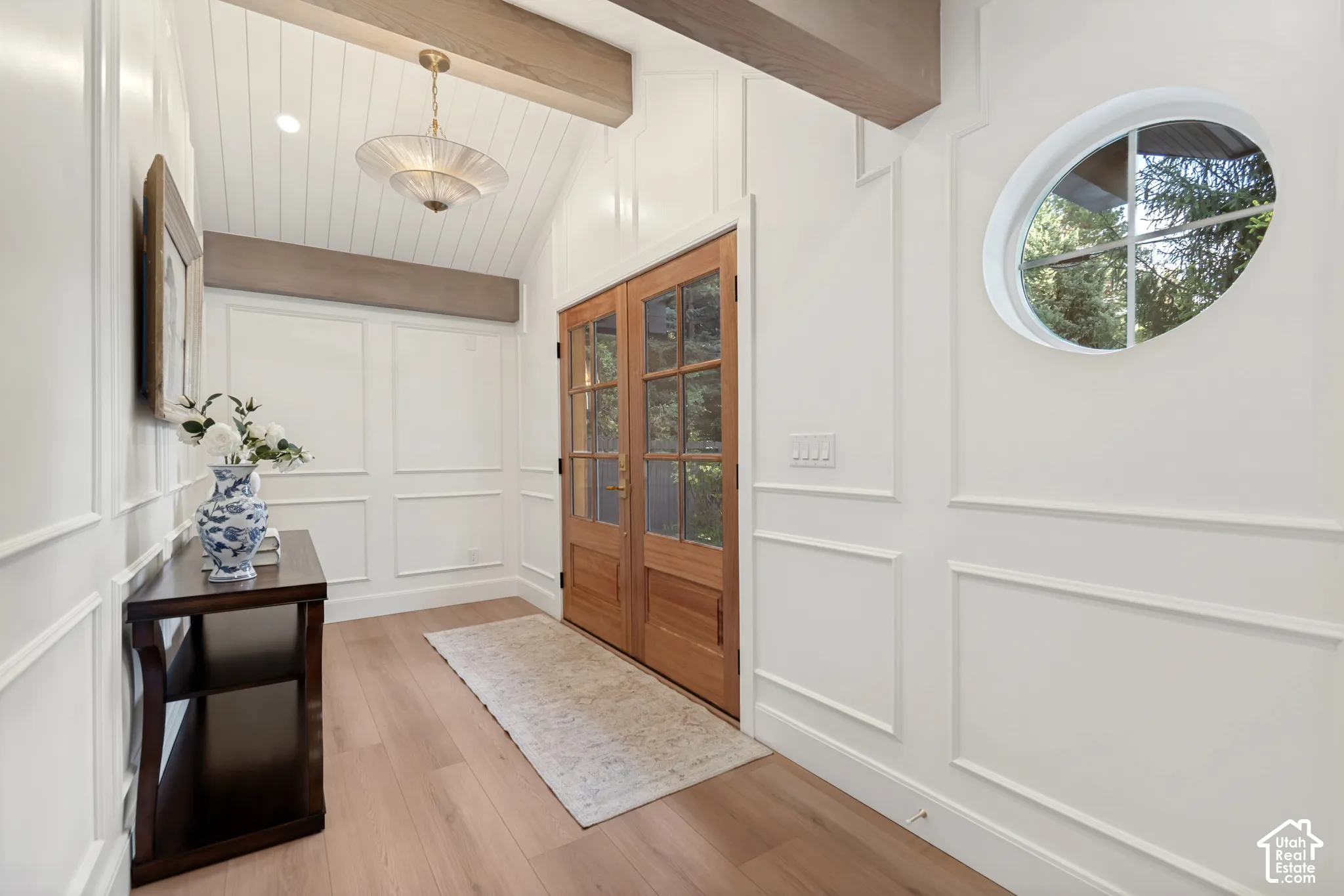 Entryway featuring light wood-finish flooring, white paneled walls, and a vaulted ceiling with exposed beams