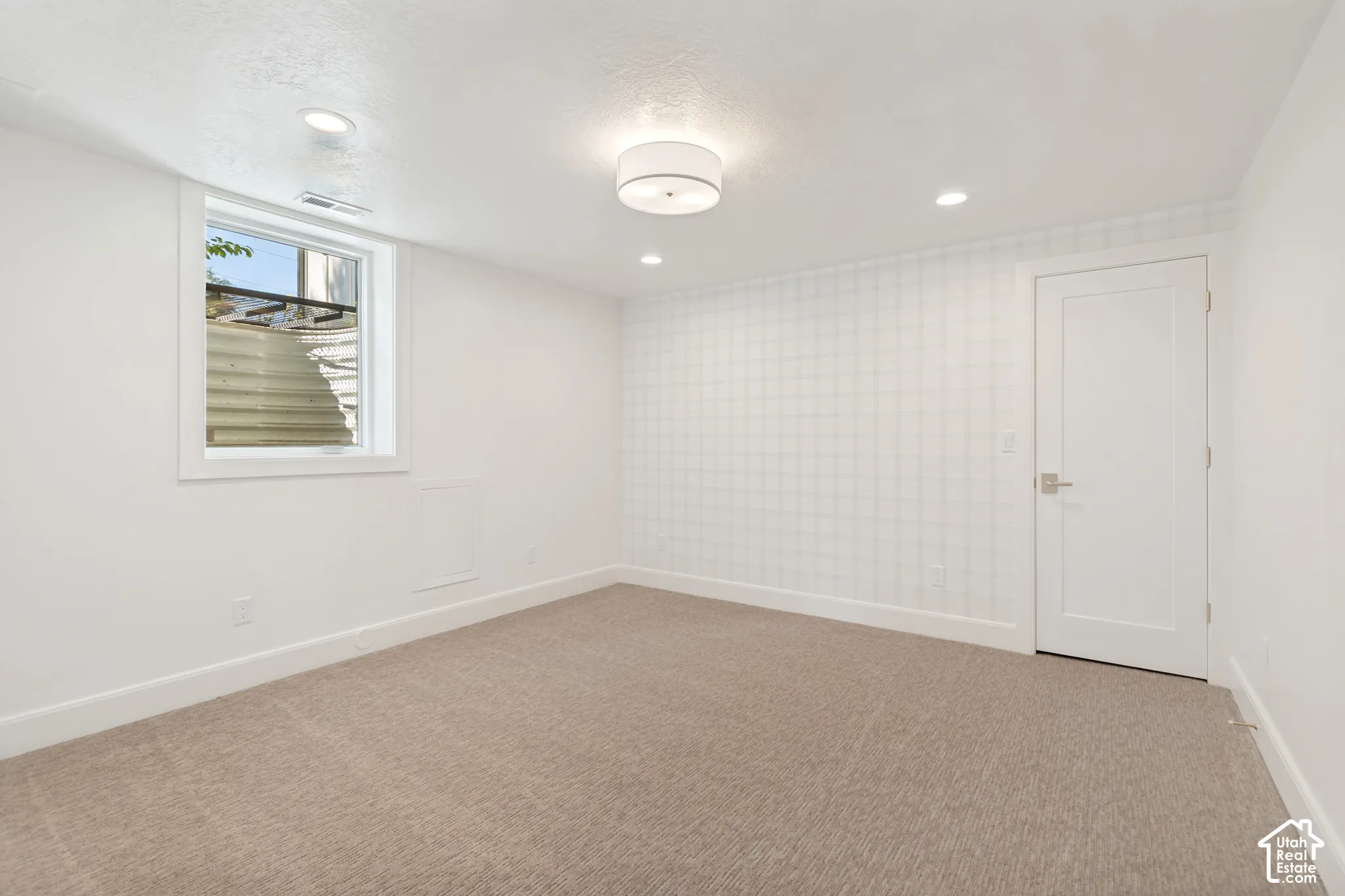 Spacious room featuring neutral carpet flooring, a single window, recessed lighting, a modern ceiling light fixture, and a solid panel door with brushed nickel hardware