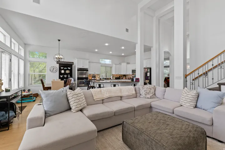 Spacious open-concept living area featuring high ceilings, wood-finish flooring, a kitchen with white cabinetry, and a staircase with wrought iron balusters