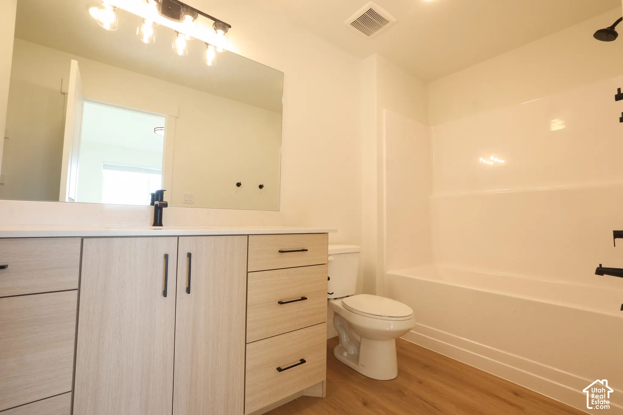 Wood-finish flooring, light-toned vanity with cabinet and drawer storage, white countertop, black fixtures, and a white bathtub with shower