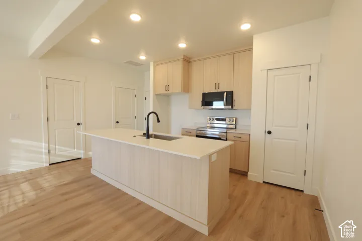 Modern kitchen featuring a central island with an integrated sink, wood-finish flooring, light-toned cabinetry, stainless steel appliances, and recessed lighting
