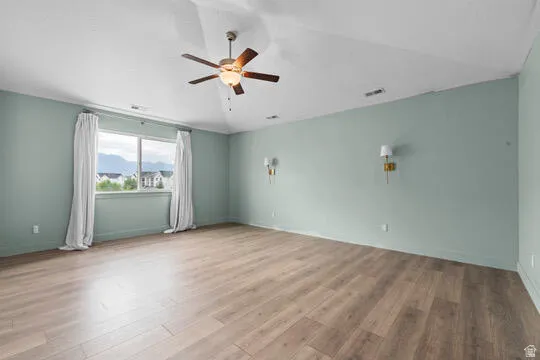 Spacious room featuring wood-finish flooring, a large window, a ceiling fan with integrated lighting, and two wall-mounted sconces