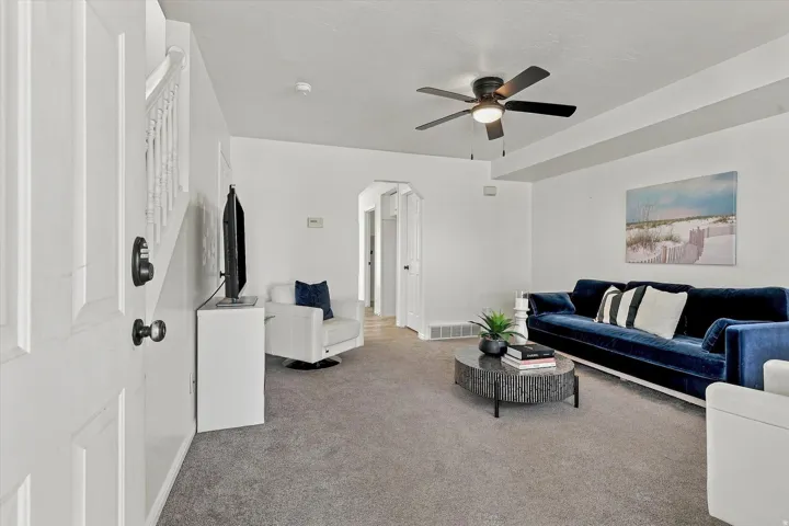 Carpeted living area featuring a ceiling fan with integrated lighting, white painted walls, white trim, and an archway leading to another room