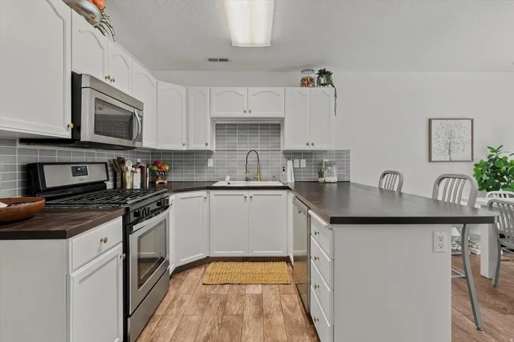 White cabinetry with gold-tone hardware, wood-finish flooring, stainless steel appliances, dark countertops, and a gray subway tile backsplash