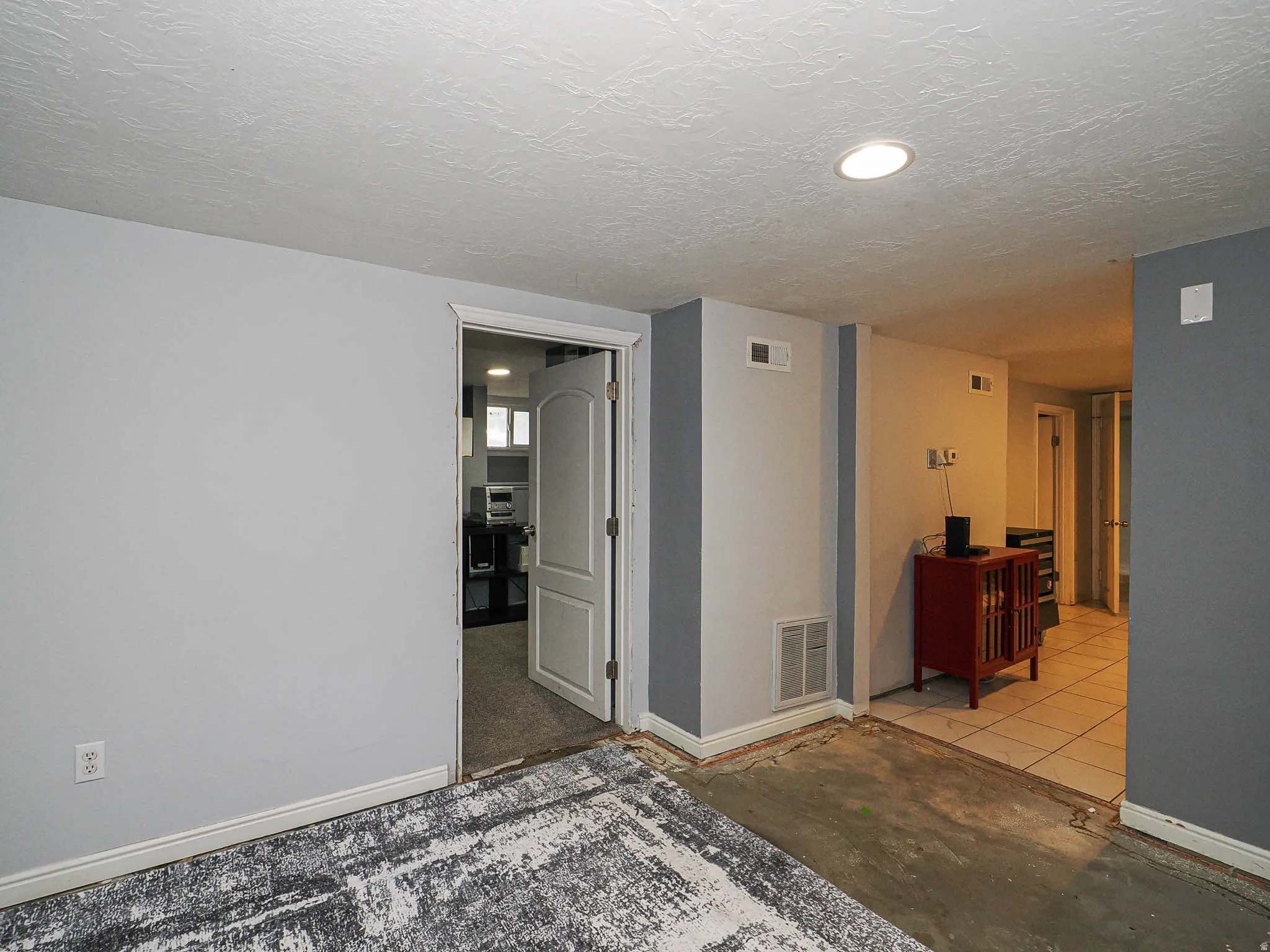 Recessed lighting illuminates a space featuring light gray walls, white baseboards, and a partial area with a patterned floor covering