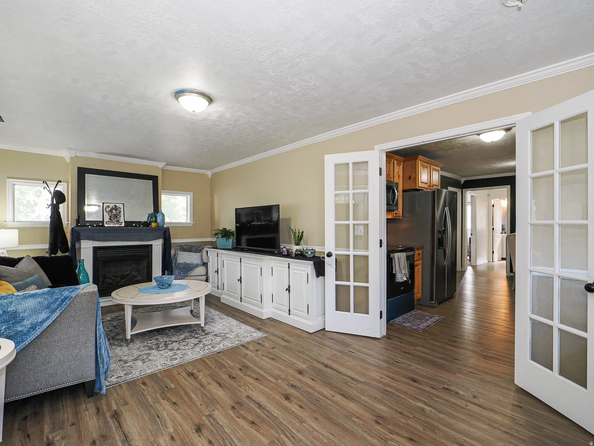 Living area featuring wood-finish flooring, a fireplace with a white mantle, two windows, crown molding, and a built-in white cabinet unit