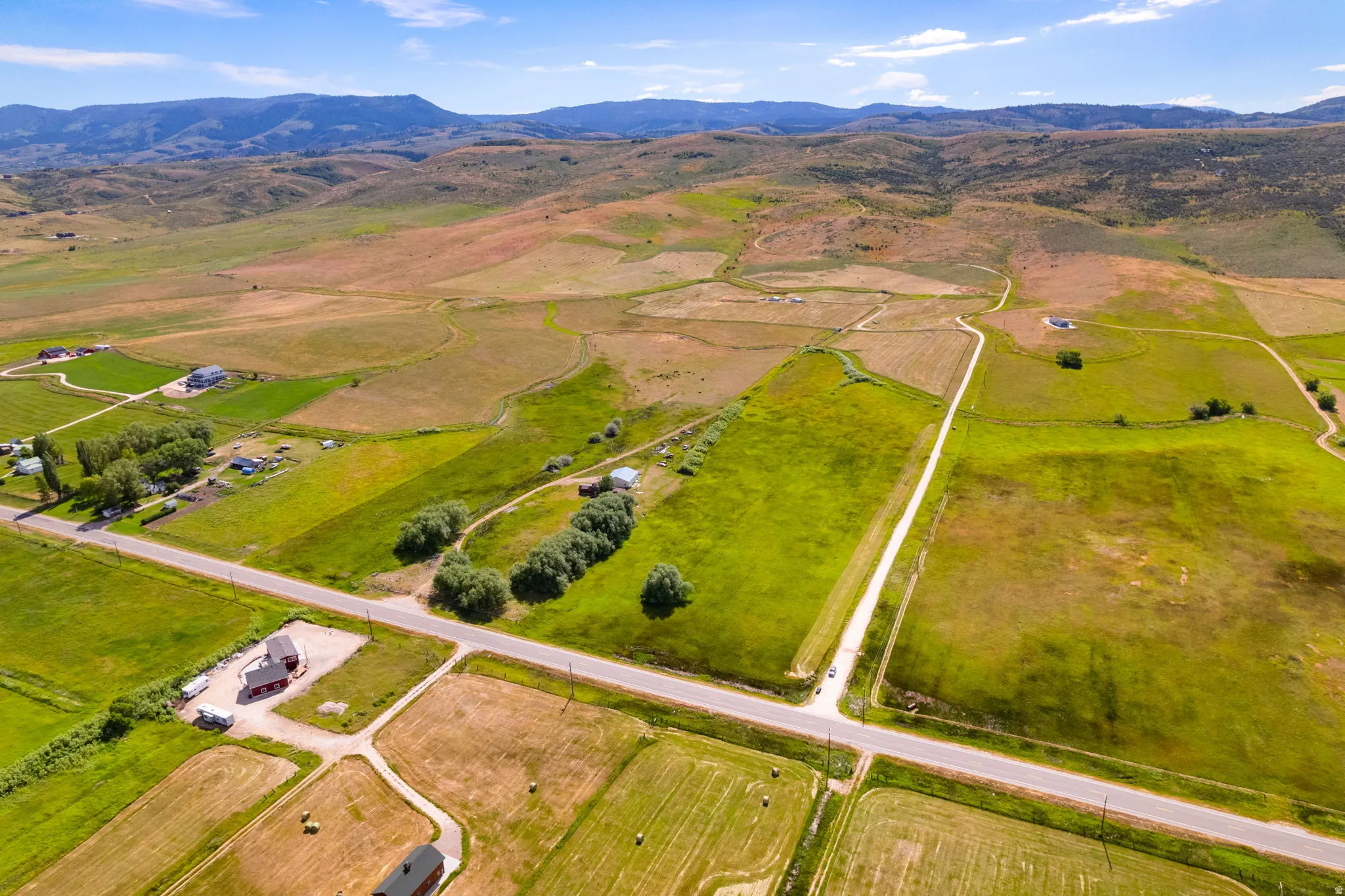 Expansive rural landscape featuring a network of roads, varied green fields, and distant mountains under a blue sky with scattered clouds