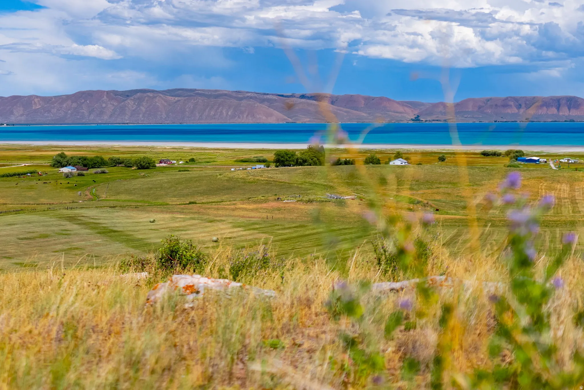 Expansive open landscape with distant water, rolling hills, and scattered structures under a partly cloudy sky