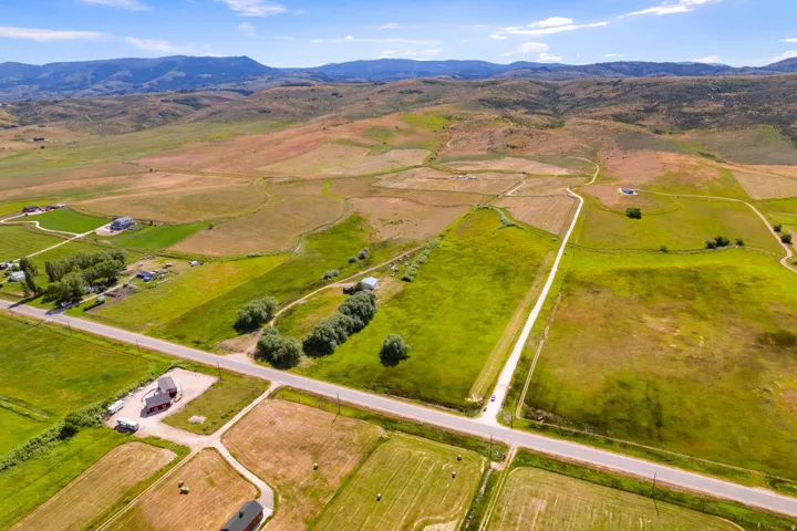 Expansive rural landscape featuring a network of roads, varied green fields, and distant mountains under a blue sky with scattered clouds