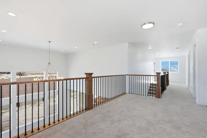 Upper-level landing featuring light-colored carpeting, a wood-finish handrail with black metal balusters, recessed lighting, and a prominent chandelier visible below