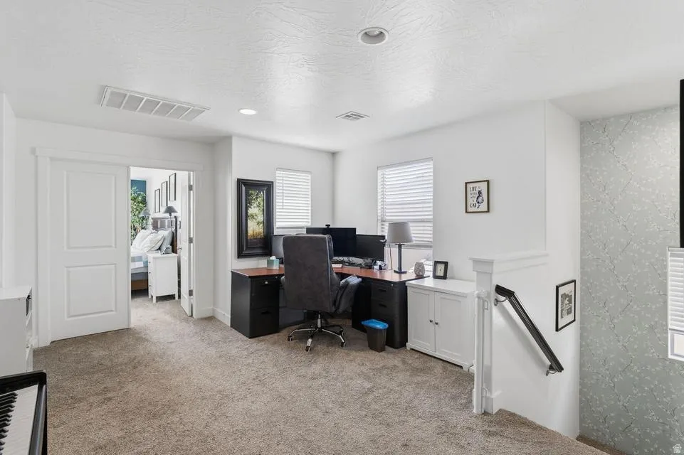 Carpeted upper-level space featuring a partial view of a bedroom through double pocket doors, a dedicated office area with a window, recessed lighting, and a staircase with a white balustrade and black handrail