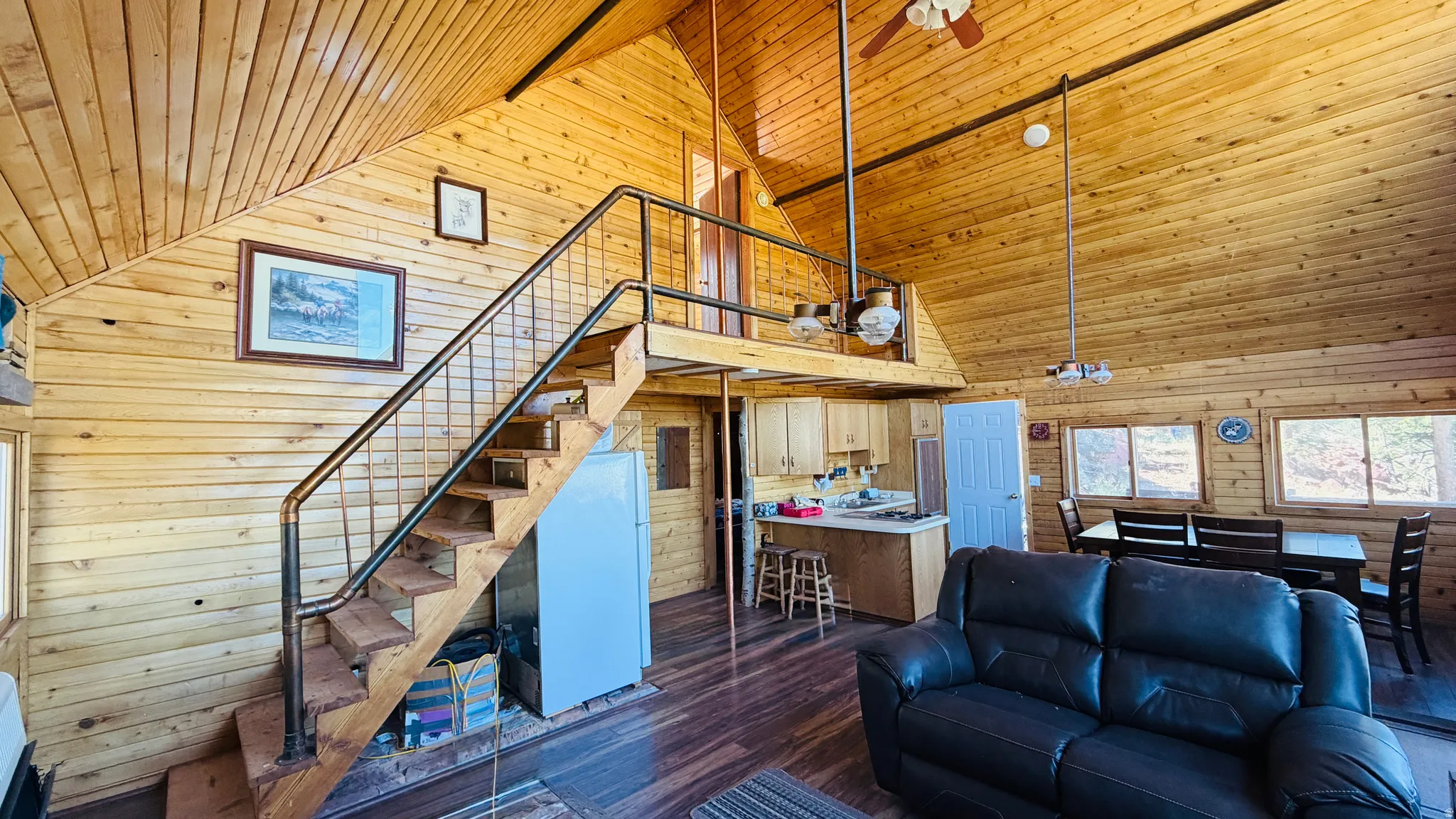 Vaulted wood-paneled ceilings with exposed beams, wood-finish flooring, a contemporary staircase with metal railing, an open kitchen area with light wood cabinetry, and multiple windows providing natural illumination
