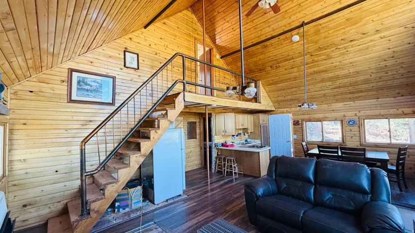 Vaulted wood-paneled ceilings with exposed beams, wood-finish flooring, a contemporary staircase with metal railing, an open kitchen area with light wood cabinetry, and multiple windows providing natural illumination