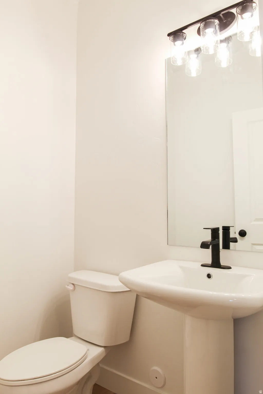 Pedestal sink with a matte black faucet, wall-mounted rectangular mirror, and a contemporary four-light vanity fixture