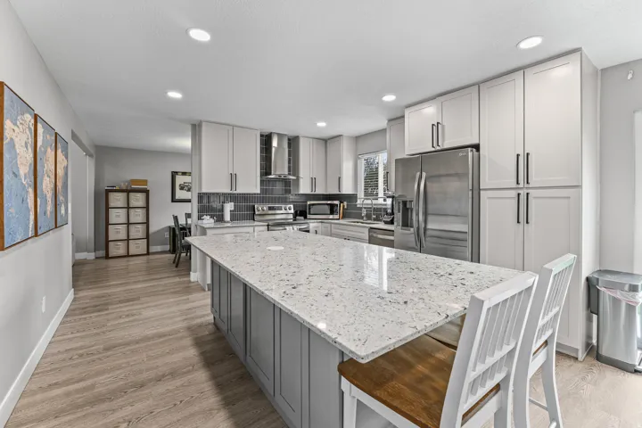 Modern kitchen featuring a large island with a light-toned countertop, light gray base cabinetry, light-toned upper cabinetry with matte black hardware, stainless steel appliances, and wood-finish flooring