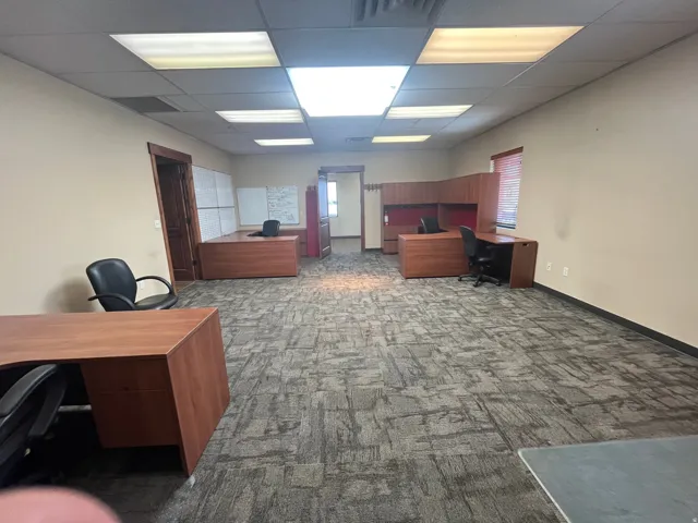 Spacious commercial office area featuring patterned carpet flooring, recessed ceiling lighting, light beige walls, and built-in wood-finish desks with shelving