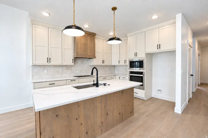 Kitchen featuring a central island with a sink, white cabinetry with black hardware, integrated stainless steel appliances, a wood-finish range hood, and light wood-finish flooring