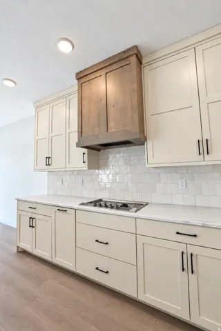 Kitchen featuring light-toned cabinetry with black hardware, an integrated gas cooktop, a wood-finish range hood, and a white subway tile backsplash