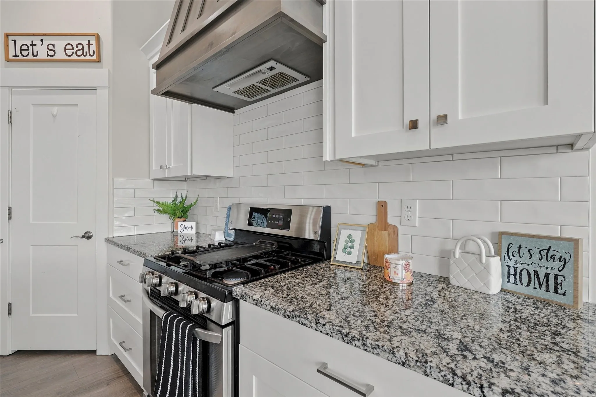 Modern kitchen featuring white shaker cabinetry, speckled countertop, subway tile backsplash, stainless steel range, and a wood-finish range hood