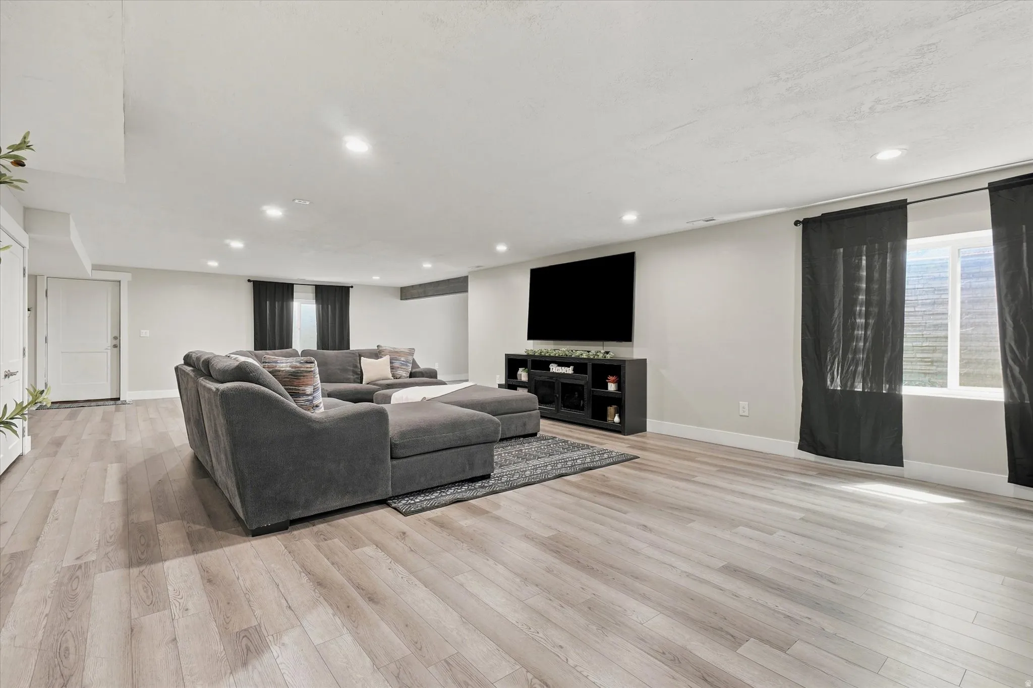 Spacious living area featuring light wood-finish flooring, recessed lighting, white walls, and a large window