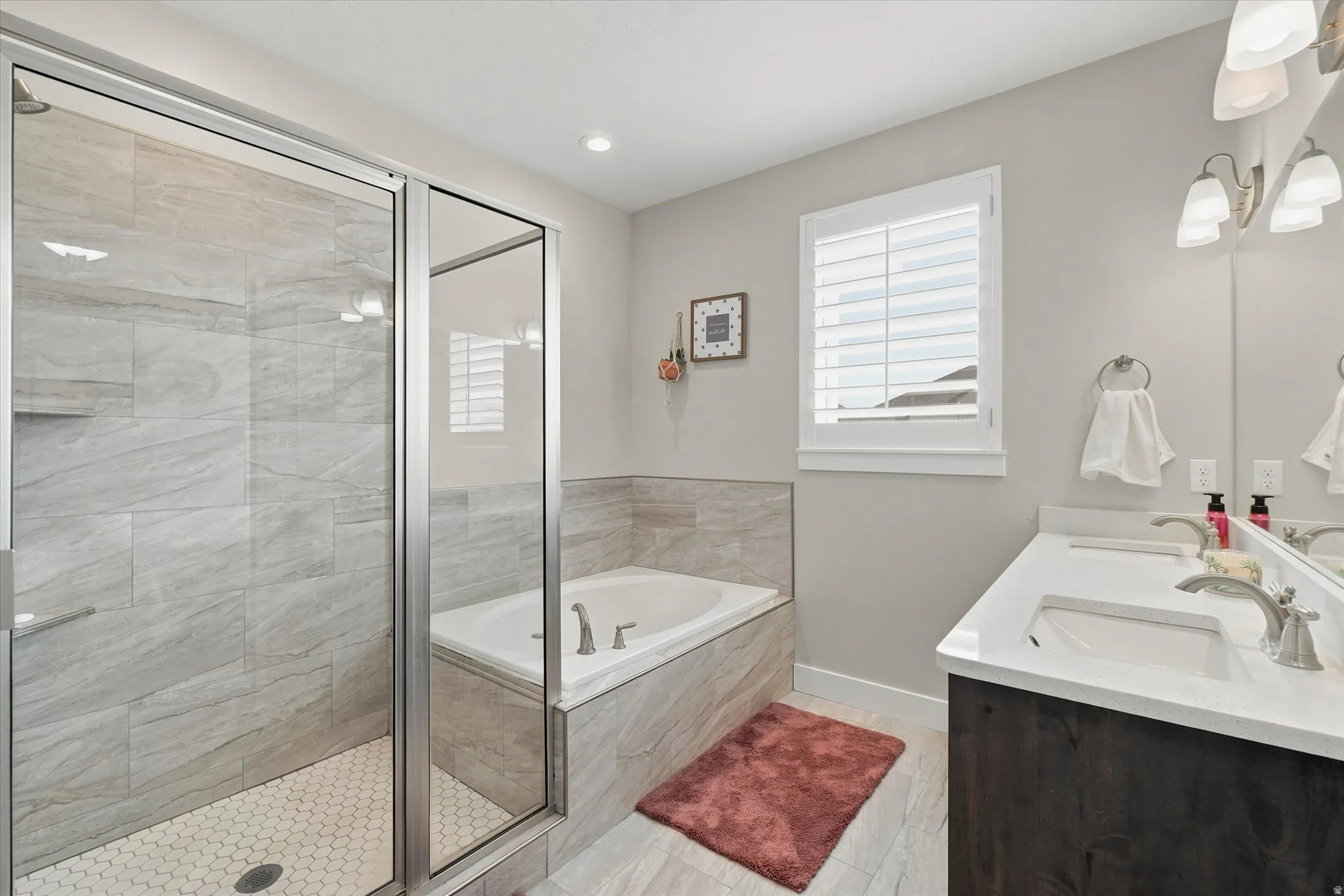 Bathroom featuring a glass-enclosed shower with large format tile surround and hexagonal tile pan, a built-in soaking tub with a tiled surround, a double vanity with an integrated countertop, dark wood-finish cabinetry, and a window with plantation shutte