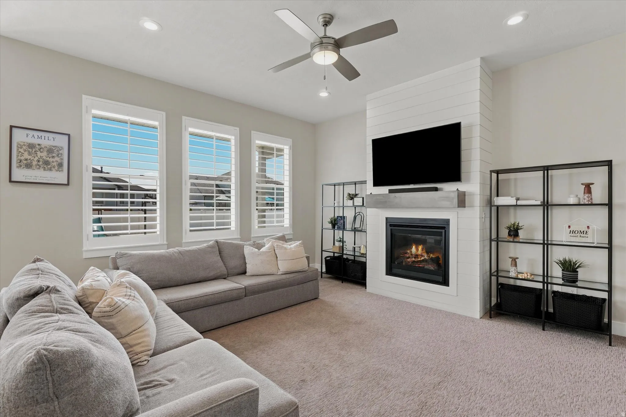 Spacious living area featuring a fireplace with a shiplap accent wall, built-in shelving, a ceiling fan, recessed lighting, and triple windows with plantation shutters