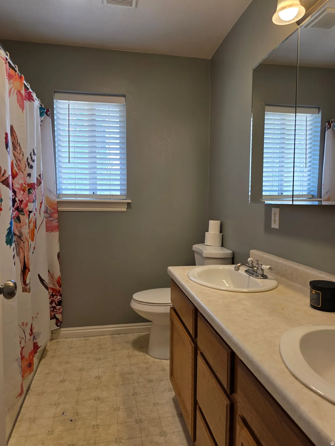 Bathroom featuring a double vanity with integrated sinks, a mirror with overhead lighting, a toilet, and a window with blinds