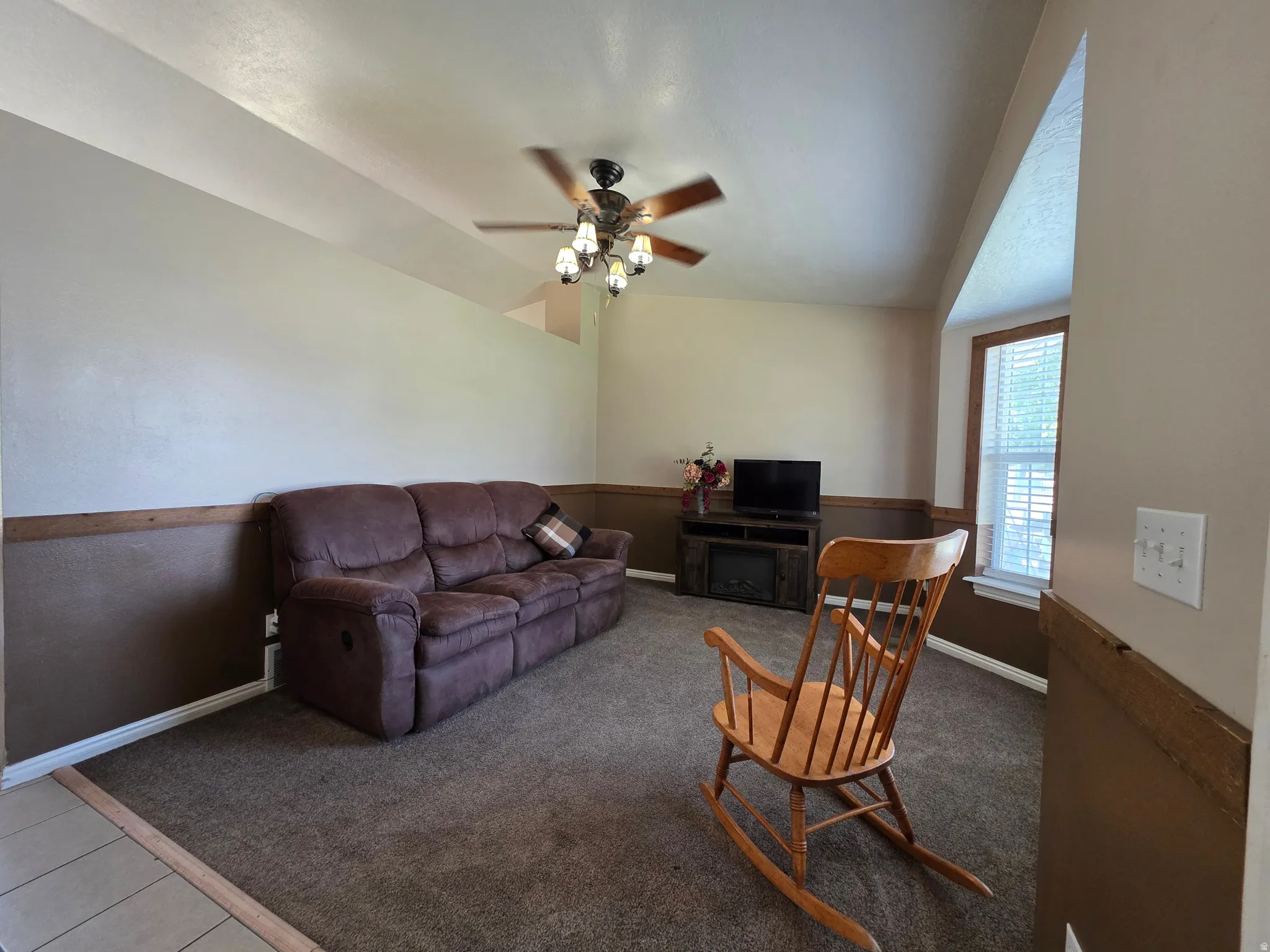 Spacious living area featuring neutral two-tone walls with wood trim, a ceiling fan with integrated lighting, a window with blinds, and plush carpet flooring