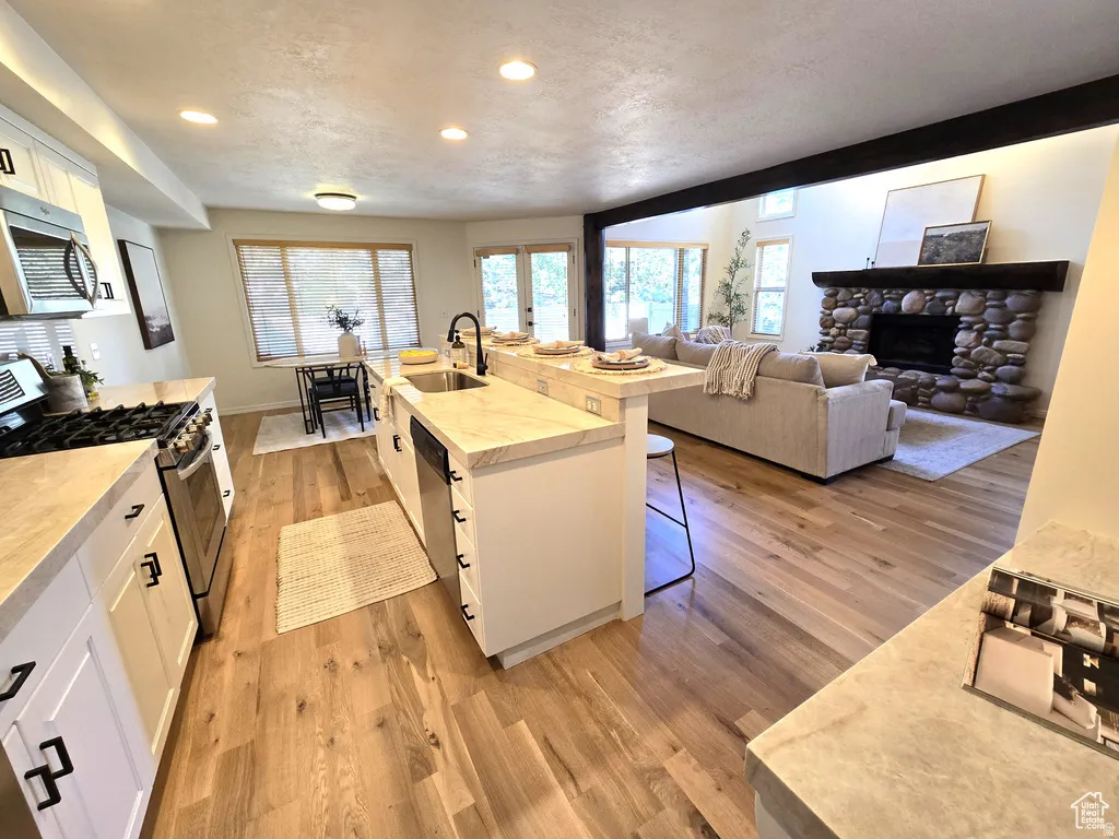 Open-concept living area featuring a kitchen island with seating, stone-faced fireplace, exposed beam ceiling, stainless steel appliances, and wood-finish flooring
