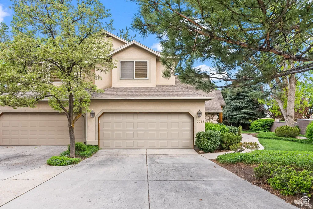 Stucco exterior featuring a two-car garage, concrete driveway, and mature landscaping