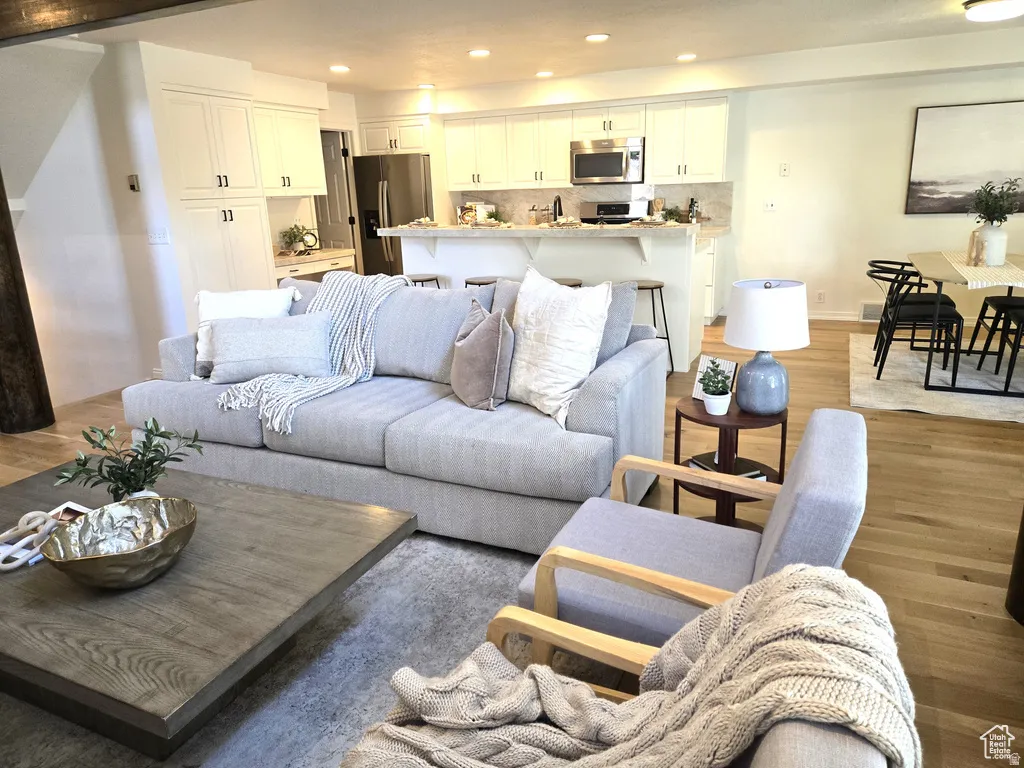 Open-concept living space featuring wood-finish flooring, recessed lighting, and a white kitchen with stainless steel appliances and a breakfast bar